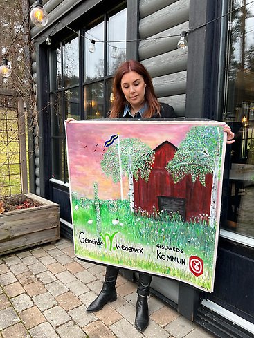 En kvinna står framför en husfasad och håller upp en målad tavla.