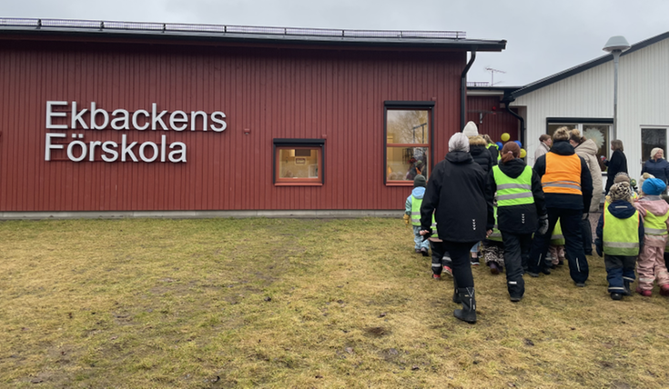 En röd byggnad med texten "Ekbackens förskola". Till höger i bild står flera vuxna och barn i reflexvästar med ryggen vänd mot kameran.