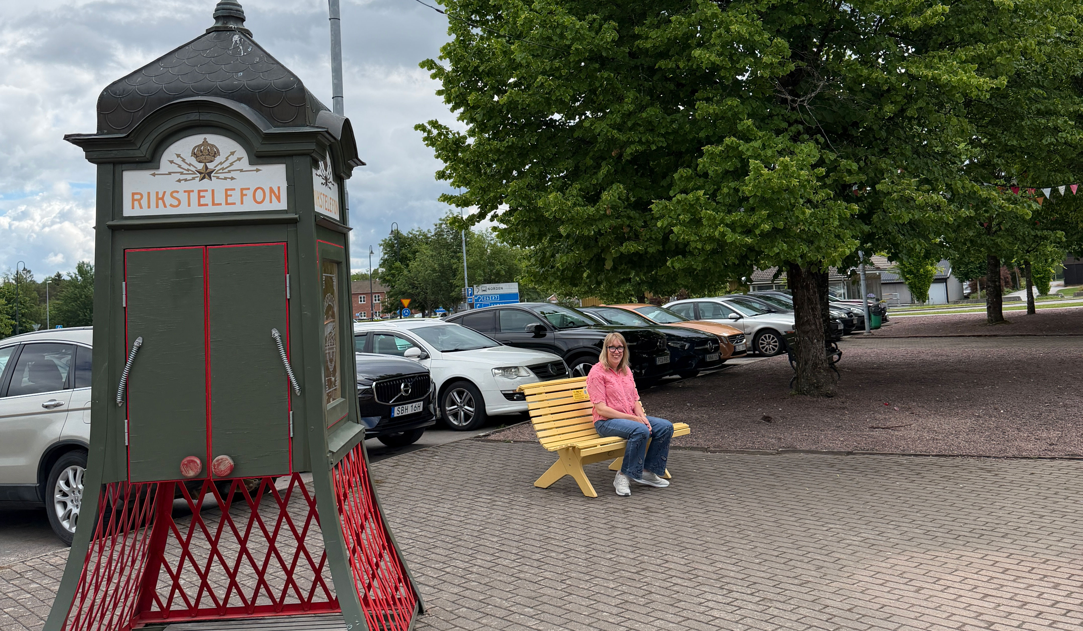 Lina sitter på en gul bänk bredvid en telefonkiosk märkt "RIKSTELEFON". Flera bilar parkerar på asfalterad plats.