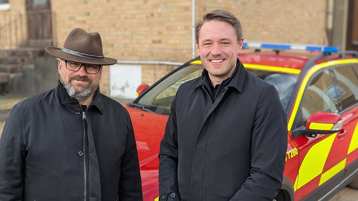 Två glada personer framför räddningstjänstens fordon.