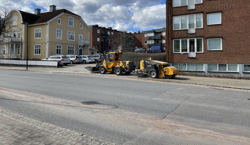 En gul sopmaskin i arbete på en cykelväg. I bakgrunden syns ett gult trähus och ett brunt tegelhus.