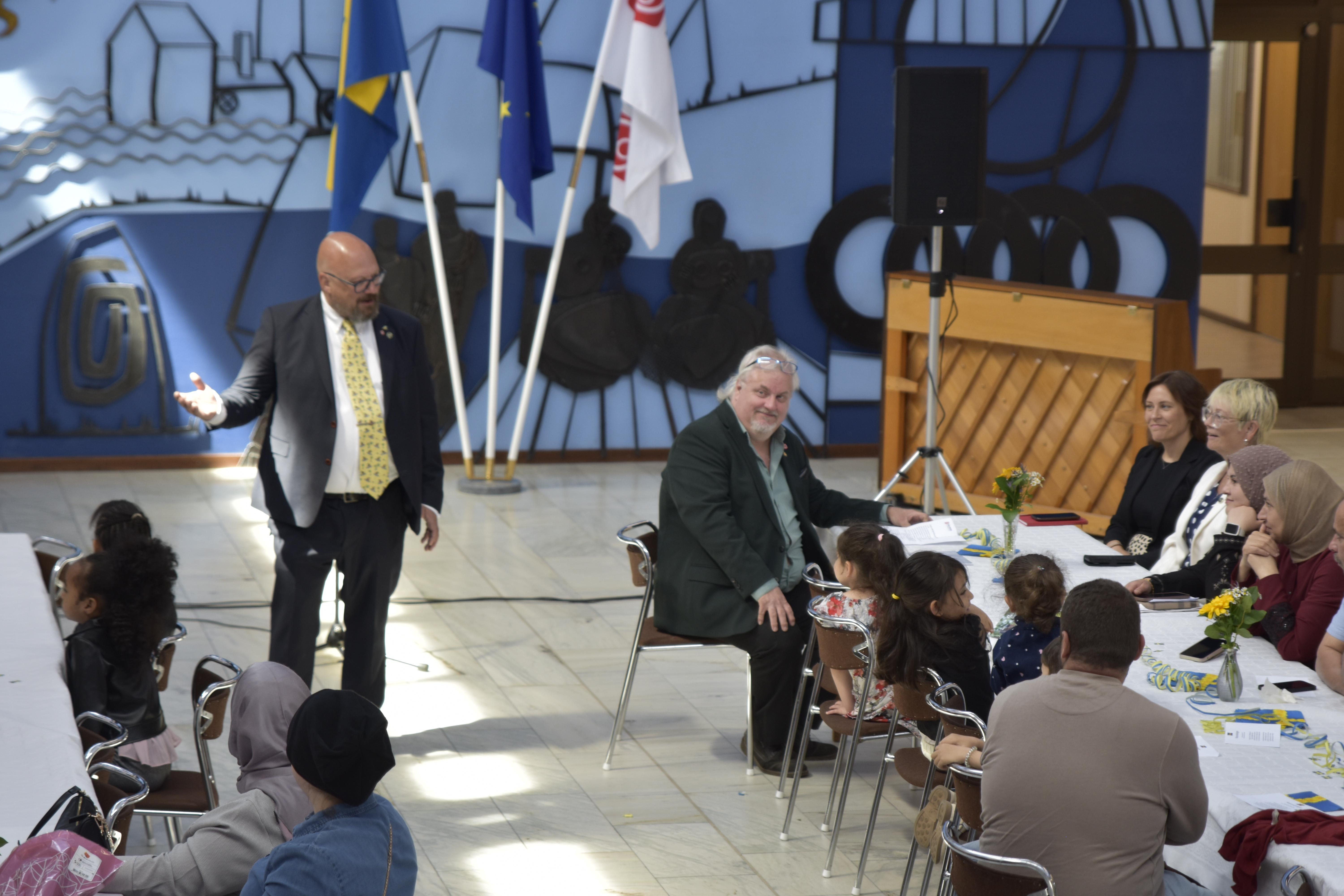 Ett tiotal personer sitter vid två långbord och tittar mot kommunfullmäktiges ordförande som håller tal.