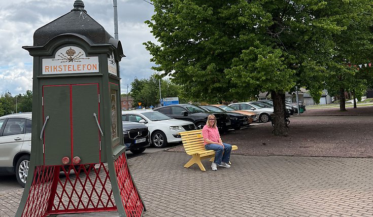 Lina sitter på en gul bänk bredvid en telefonkiosk märkt "RIKSTELEFON". Flera bilar parkerar på asfalterad plats.