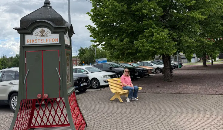 Lina sitter på en gul bänk bredvid en telefonkiosk märkt "RIKSTELEFON". Flera bilar parkerar på asfalterad plats.