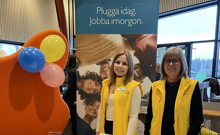 Två kvinnor i gula västar står vid ett mässbord.