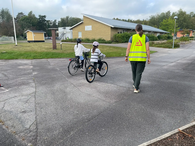 Två barn cyklar medan en pedagog i reflexväst går bredvid. 