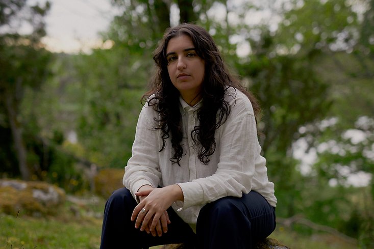 en kvinna med långt, lockigt hår sitter på huk i naturen.