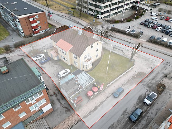 En inzoomad drönarbild som visar fastigheten Krabban 11 med omgivande gator och byggnader, samt planområdet utmarkerat med gränslinje i rött. 