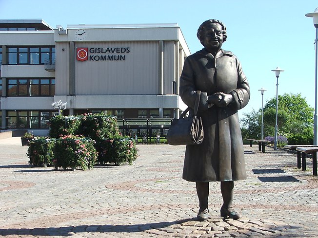 skulptur i form av en tant med handväska på ett torg framför ett grått, stort hus