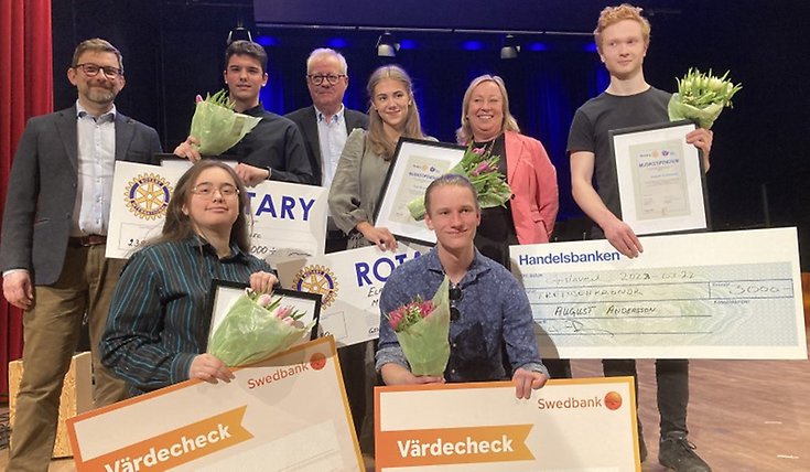 6 elever med checkar och blommor står på en scen tillsammans med representanter från Rotary. 