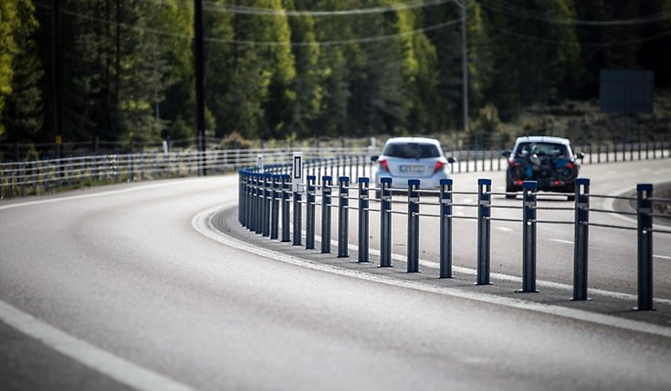 Motorväg med mitträcke. Till höger i bild syns en vit och en svart bil. 