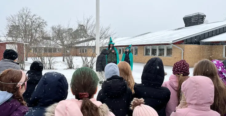 Foto på en snöig skolgård. Elever står runt flaggstången där två utklädda pedagoger står.