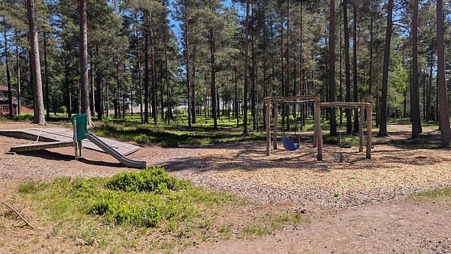 En lekplats med en rutschbana och gungor i en skogsomgivning. Marken är täckt med barkspån.
