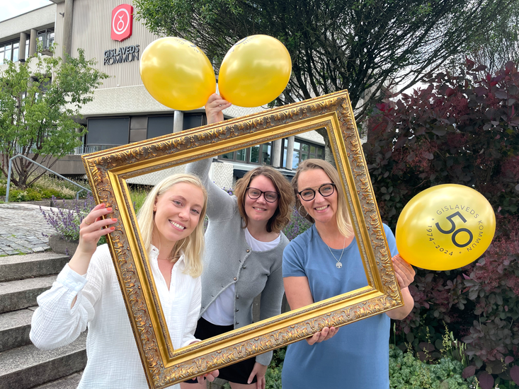 Tre kvinnor tittar fram bakom en guldig ram med ballonger i händerna. 