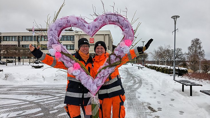 Två kvinnor i orangea arbetskläder kikar fram genom ett hjärta i rosa tyll.